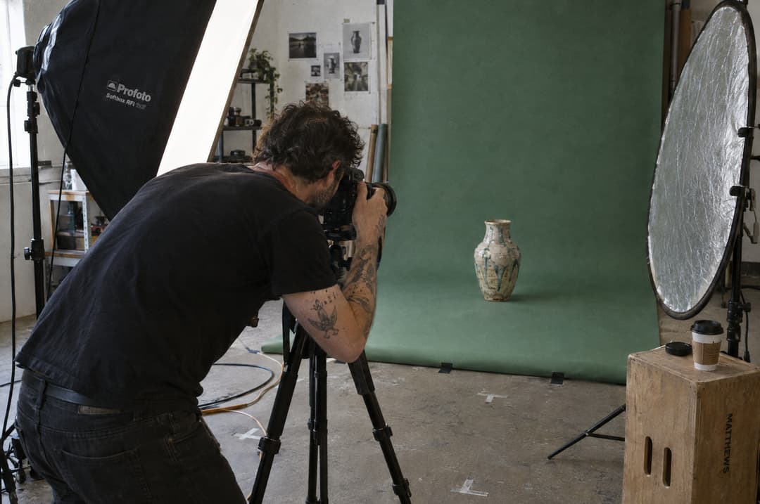 A photographer shooting a handmade ceramic vase on a green paper backdrop in a small studio