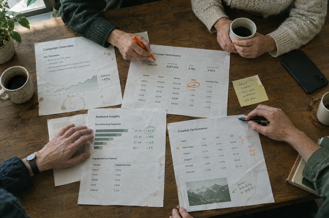 A team reviewing printed campaign results on a large wooden table, one person circling a number in orange marker